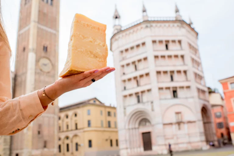 Holding a piece of Parmesan cheese on the Parma main square background in Italy. Parmesan is produced mostly in the province of Parma and was named after that producing region